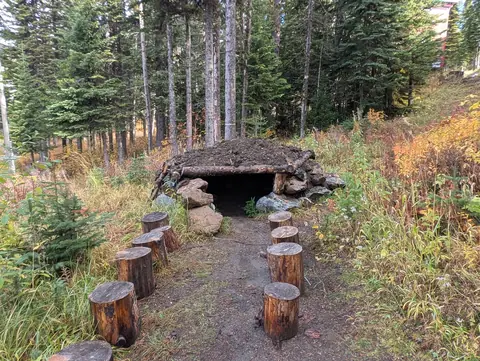 Sod-roofed log and stone bear den at the end of a path lined with two rows of log stump seats