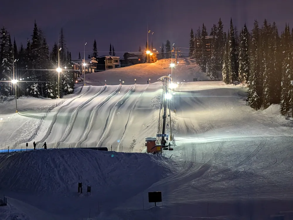 Freshly groomed ski run lit by floodlights at night, lined with snowy trees
