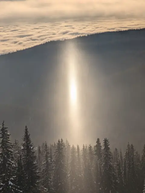 Bright column of light rising over a snowy forest and cloud-filled valley, January 26, 2025, morning