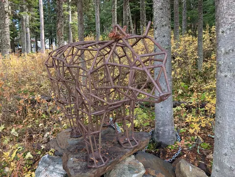 Open-frame rusty metal bear sculpture on a rock cairn among birch trunks